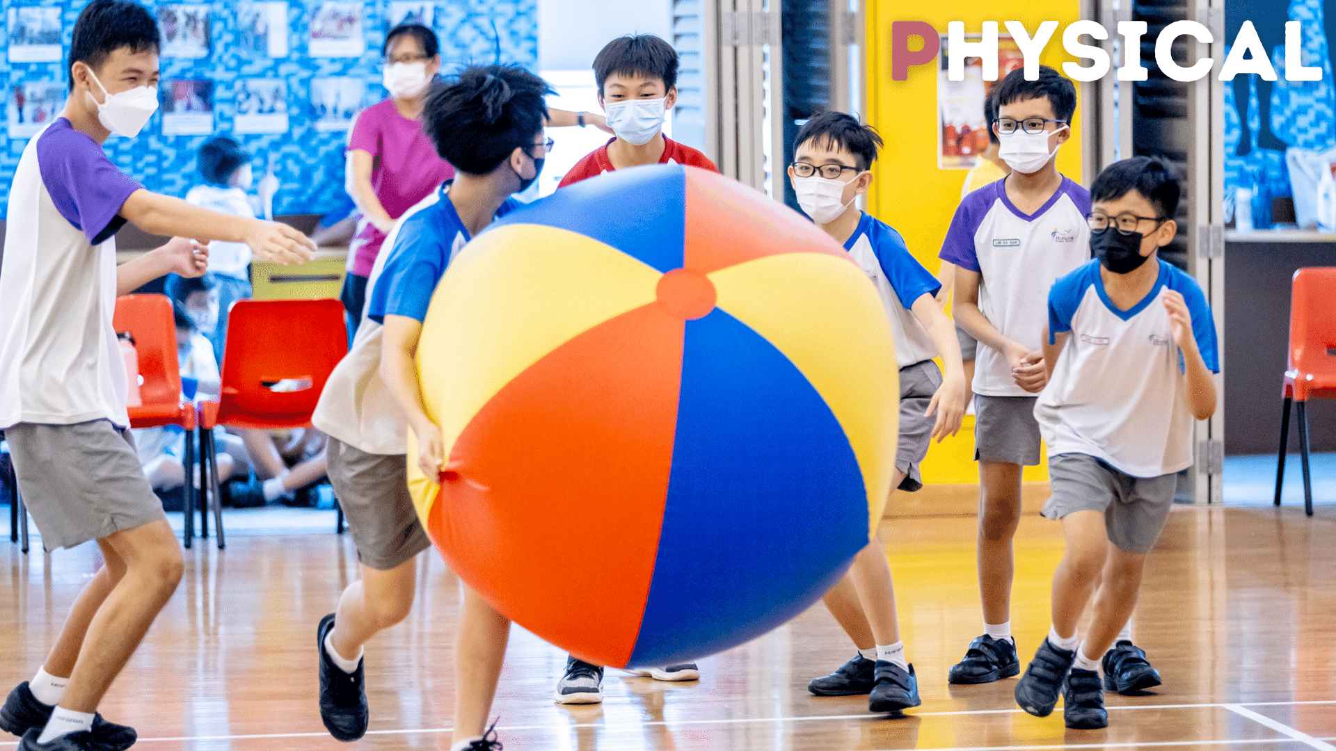 Students wearing masks play with a large colorful beach ball in a school gymnasium.